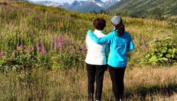 grown daughter with arm around mother's shoulder looking out into mountain scenery