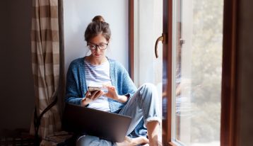 young woman in window nook using phone and laptop - time to enter recovery