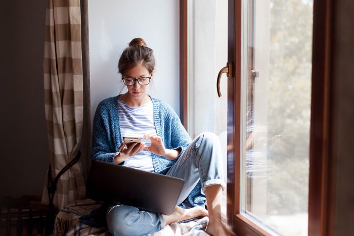 young woman in window nook using phone and laptop - time to enter recovery