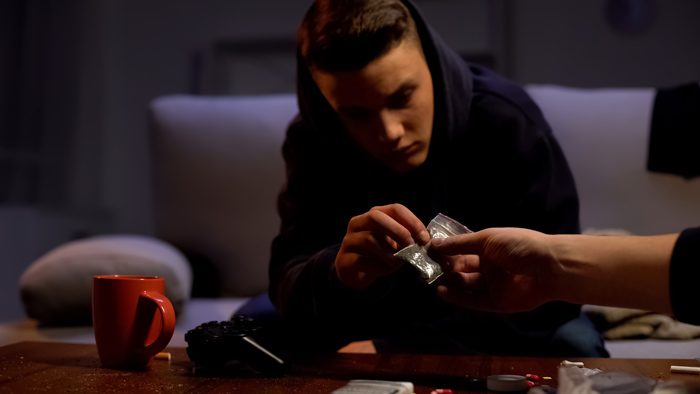 young man sitting in dark apartment with drugs and alcohol on coffee table - polysubstance misuse