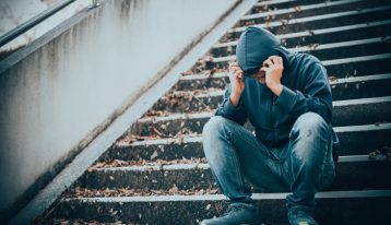 man sitting in hooded sweatshirt on stairs outdoors, visibly upset - spouse addicted