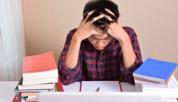 male college student at desk with hands in his hair looking stressed - stress on college students