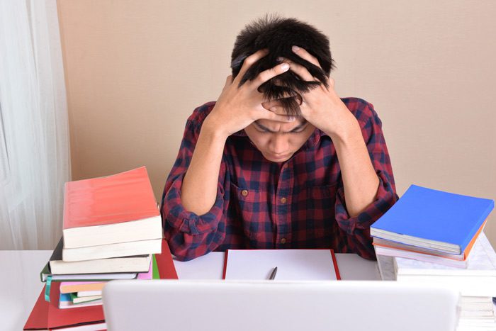 male college student at desk with hands in his hair looking stressed - stress on college students