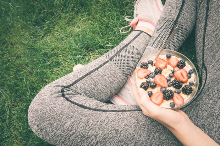 cropped shot of a woman's hands holding a bowl of oatmeal with strawberries and blueberries while sitting cross-legged outdoors in the grass; she is wearing workout pants and shoes - self-care