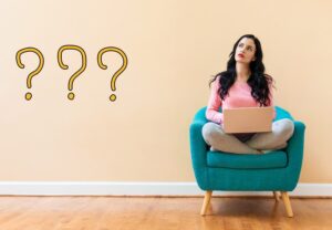 pretty brunette in pink sweater sitting in a teal blue chair and using a laptop has questioning look on her face and question marks out beside her - partial care addiction treatment