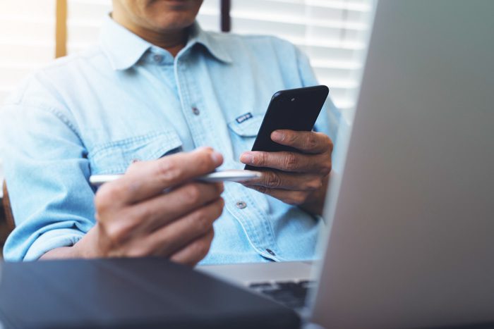 admissions, admissions specialist, admissions; cropped shot of man sitting in front of laptop and using a cell phone with one hand and has a pen in the other - what to expect