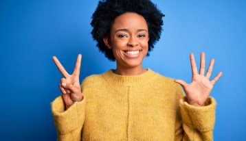 beautiful smiling young Black woman holding her hands up to sign the number seven; she is wearing a mustard colored sweater and the background is a solid medium blue - loved one's addiction