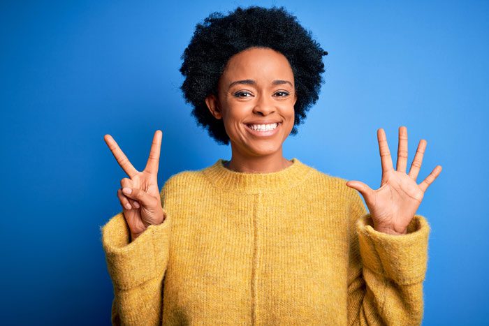 beautiful smiling young Black woman holding her hands up to sign the number seven; she is wearing a mustard colored sweater and the background is a solid medium blue - loved one's addiction