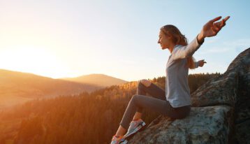 outdoor holistic addiction treatment - young woman hiker sits on edge of cliff against background of sunrise over valley. Woman is meditation and greeting a sun