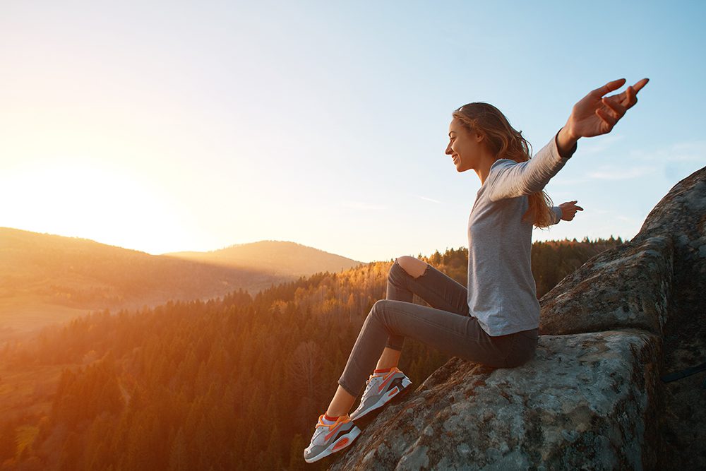 outdoor holistic addiction treatment - young woman hiker sits on edge of cliff against background of sunrise over valley. Woman is meditation and greeting a sun