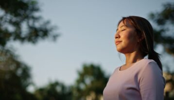 Woman standing outdoors with eyes closed, calmly taking a deep breath.