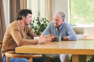 Two men sitting at a table having a serious conversation over coffee, one gesturing with his hands while the other listens attentively.