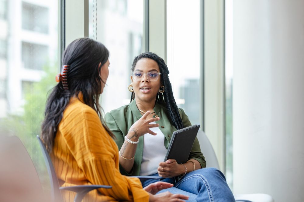 Two women sitting and talking near a large window, with one holding a tablet and actively listening, suggesting a counseling or intake session.