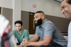 Three men sit together in a support group setting, engaged in conversation. One man in a gray shirt speaks while the others listen and smile. The group appears to be in a bright, comfortable room, fostering a sense of connection and openness.