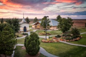 Ranch at Dove Tree, facility with landscaped gardens, walking paths, and buildings surrounded by trees under a colorful sunset sky.