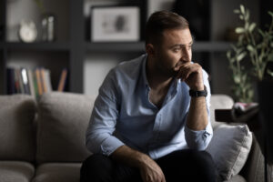 Man sitting on a couch with a serious, contemplative expression, resting his hand on his chin, suggesting emotional struggle or reflection.