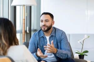 Man engaged in a therapy session, speaking thoughtfully while sitting across from another person in a bright, comfortable office.