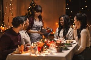 A group of friends sit around a festive holiday table as a woman brings a roasted dish to serve. Warm lights, candles, and seasonal décor create a cozy, joyful atmosphere.