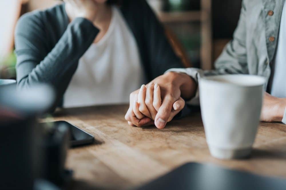 A close-up of two people sitting at a table, gently holding hands in a supportive and comforting gesture.