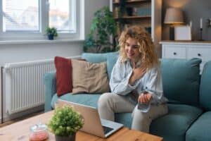 A woman with curly hair sits on a blue sofa, smiling at a laptop on a wooden table. She is engaged is a telehealth appointment.